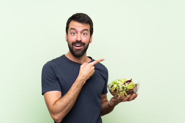 Giovane uomo bello con insalata sopra la parete verde isolata sorpresa e lato indicante