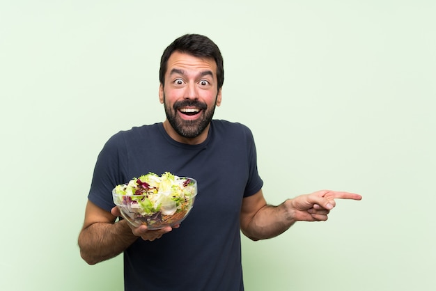 Giovane uomo bello con insalata sorpresa e puntando il dito verso il lato