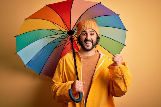 Giovane uomo bello con la barba che indossa un impermeabile per i giorni di pioggia con un ombrello colorato che urla orgoglioso e celebra la vittoria e il successo molto eccitato emozione applaudente