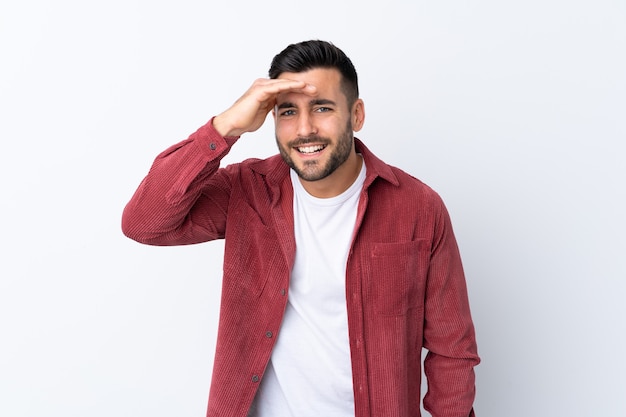 Giovane uomo bello con la barba che indossa una giacca di velluto a coste su sfondo bianco isolato che guarda lontano con la mano per guardare qualcosa