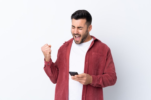 Giovane uomo bello con la barba che indossa una giacca di velluto a coste su sfondo bianco isolato con il telefono in posizione di vittoria