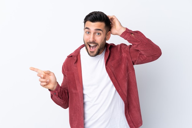 Giovane uomo bello con la barba che indossa una giacca di velluto a coste su sfondo bianco isolato sorpreso e puntare il dito di lato