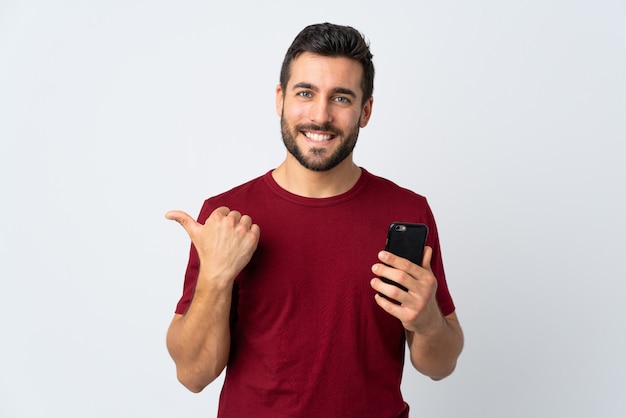 Giovane uomo bello con la barba che per mezzo del telefono cellulare isolato sulla parete bianca che indica il lato per presentare un prodotto