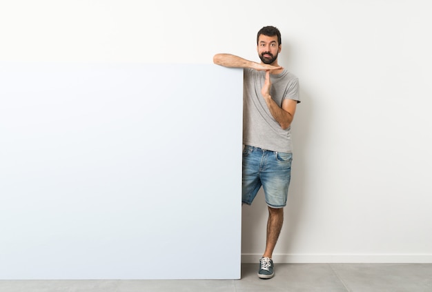 Giovane uomo bello con la barba che tiene un grande cartello vuoto blu che fa gesto di time out