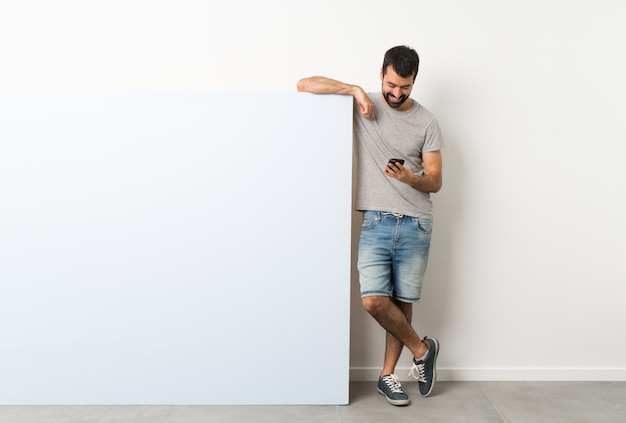 Giovane uomo bello con la barba che tiene un grande cartello vuoto blu che invia un messaggio con il cellulare