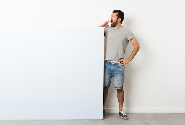 Giovane uomo bello con la barba che tiene un grande cartello vuoto blu che sbadiglia e che copre la bocca spalancata con la mano