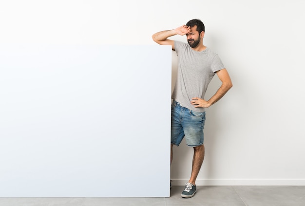 Giovane uomo bello con la barba che tiene un grande cartello vuoto blu con espressione stanca e malata