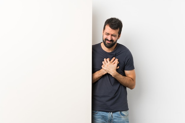 Giovane uomo bello con la barba che tiene un grande cartello vuoto che ha un dolore nel cuore