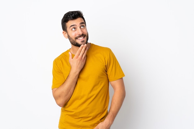 Giovane uomo bello con la barba isolata sulla parete bianca che osserva in su mentre sorride