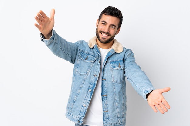 Giovane uomo bello con la barba isolata sulla parete bianca che presenta e che invita a venire con la mano