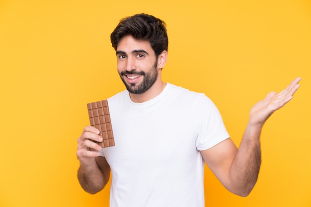 Giovane uomo bello con la barba sopra il muro giallo isolato prendendo una tavoletta di cioccolato e sorpreso