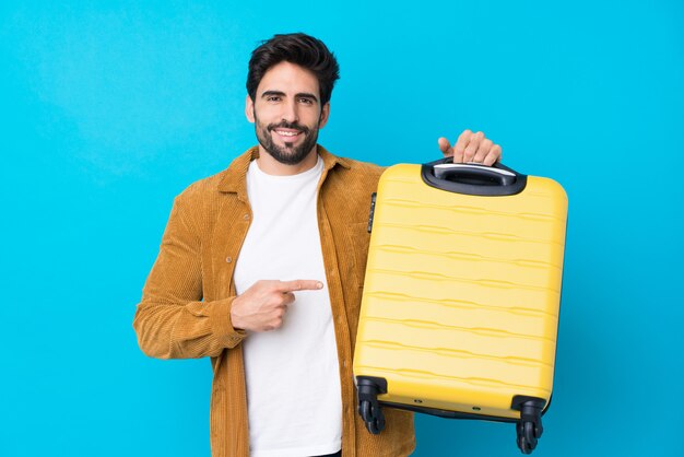 Giovane uomo bello con la barba sopra la parete blu isolata in vacanza con la valigia di viaggio