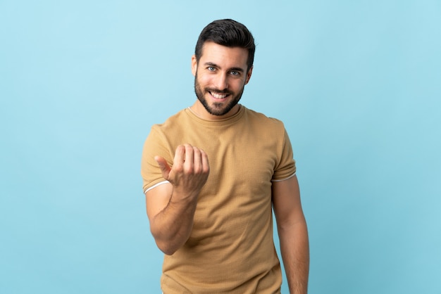 Giovane uomo bello con la barba sopra la parete isolata che invita a venire con la mano. Felice che tu sia venuto