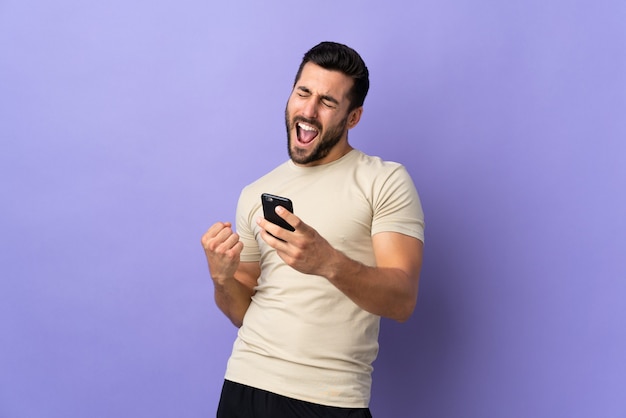 Giovane uomo bello con la barba sopra la parete isolata con il telefono in posizione di vittoria