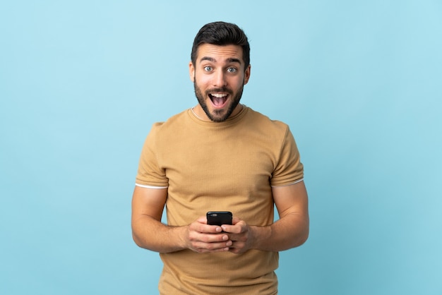 Giovane uomo bello con la barba sopra la parete isolata sorpreso e l'invio di un messaggio