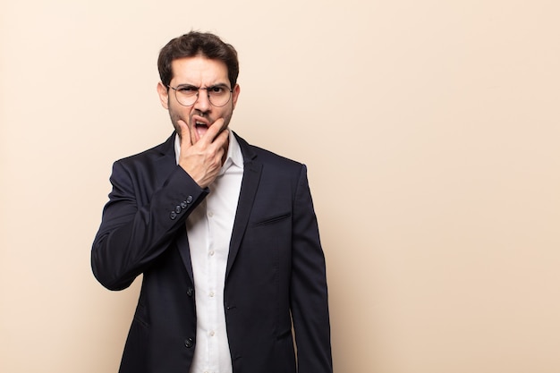 Giovane uomo bello con la bocca e gli occhi spalancati e la mano sul mento, sentendosi spiacevolmente scioccato, dicendo cosa o wow