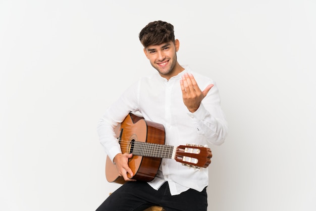 Giovane uomo bello con la chitarra sopra isolato bianco che invita a venire con la mano. Felice che tu sia venuto