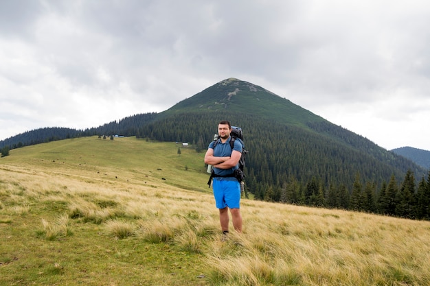 Giovane uomo bello con lo zaino che sta nella valle erbosa della montagna