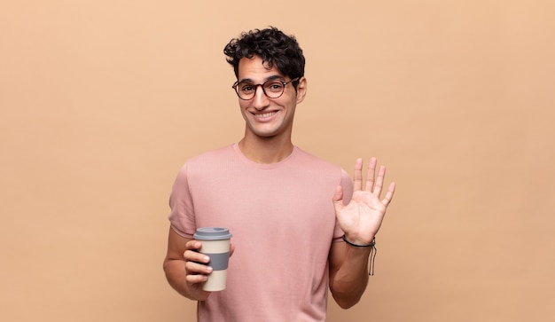 Giovane uomo bello con un caffè che sorride allegramente e allegramente, agitando la mano, dandoti il benvenuto e salutandoti o salutandoti