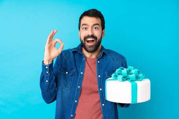 Giovane uomo bello con una grande torta sopra l'azzurro sorpreso e mostrando il segno giusto
