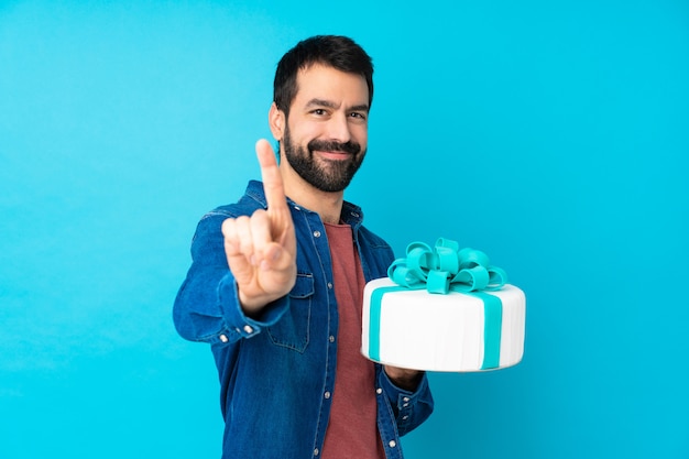 Giovane uomo bello con una grande torta sopra la parete blu isolata che mostra e che solleva un dito