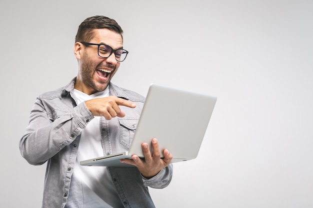 Giovane uomo bello divertente in computer portatile della tenuta della camicia e sorridere mentre stando contro il fondo bianco. Divertiti.