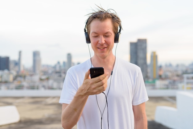 Giovane uomo bello felice con capelli biondi che per mezzo del telefono mentre ascoltando la musica contro la vista della città