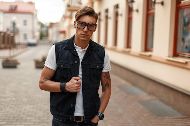 Giovane uomo bello in occhiali alla moda in gilet di jeans vintage e una camicia bianca vicino all'edificio