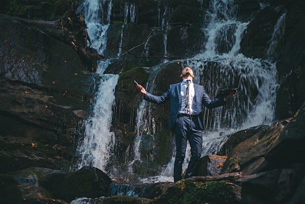 Giovane uomo bello in tuta da ufficio bagnata che tiene il computer portatile sotto l'acqua sullo sfondo della natura. Fantasia sul resto. Burnout nel concetto di lavoro.