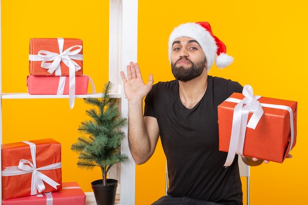 Giovane uomo bello in un cappello di Babbo Natale è in possesso di regali di congratulazioni accanto a un albero di Natale