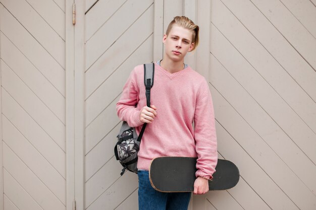 Giovane uomo bello in un maglione rosa con uno skateboard e uno zaino vicino a una parete di legno