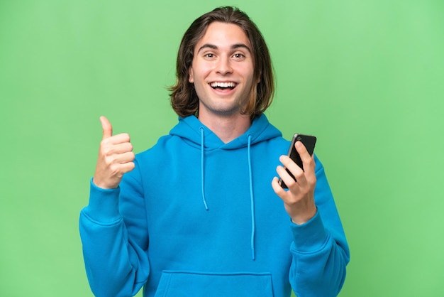 Giovane uomo bello isolato su sfondo verde croma utilizzando il telefono cellulare mentre si fa il pollice in alto