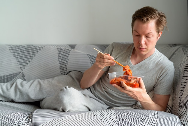 Giovane uomo bello mangiare kimchi sdraiato sul divano di casa