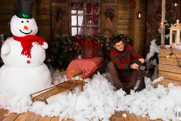 Giovane uomo bello seduto su un decoro di neve in cotone sul pavimento mentre tiene i pattini da ghiaccio in una casa in legno decorata con un grande pupazzo di neve invernale.