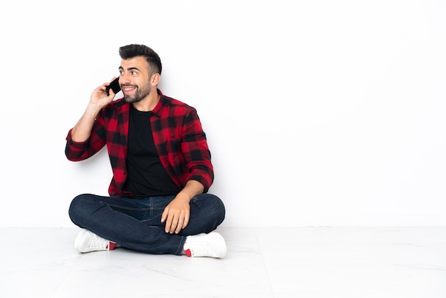 Giovane uomo bello seduto sul pavimento mantenendo una conversazione con il telefono cellulare