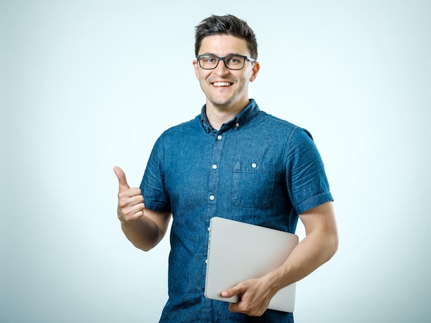 Giovane uomo bello sicuro in camicia che tiene il computer portatile mentre stando contro il fondo bianco