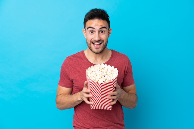 Giovane uomo bello sopra la parete blu isolata che tiene un grande secchio di popcorn