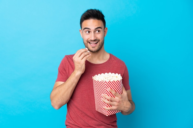 Giovane uomo bello sopra la parete blu isolata che tiene un grande secchio di popcorn