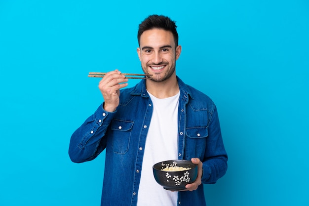 Giovane uomo bello sopra la parete blu isolata che tiene una ciotola di tagliatelle con le bacchette