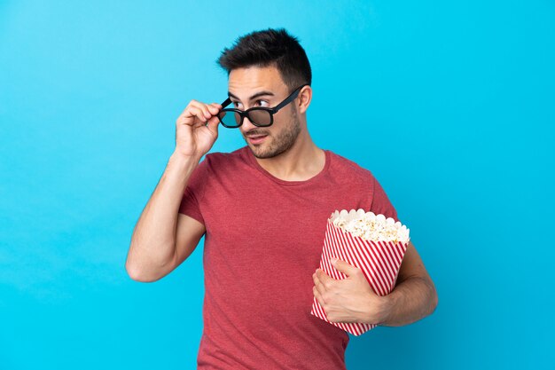 Giovane uomo bello sopra la parete blu isolata con gli occhiali 3d e tenere un grande secchio di popcorn