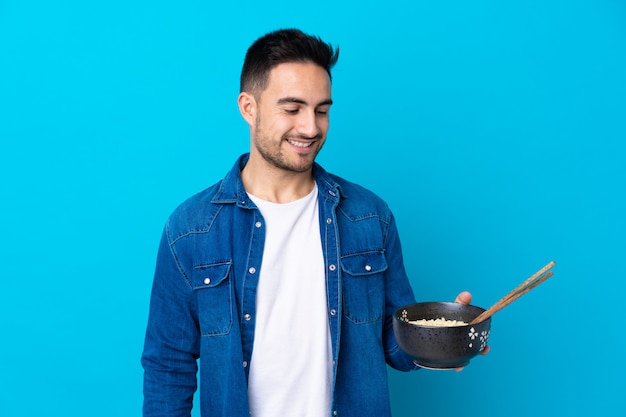 Giovane uomo bello sopra la parete blu isolata con l'espressione felice mentre tenendo una ciotola di tagliatelle con le bacchette