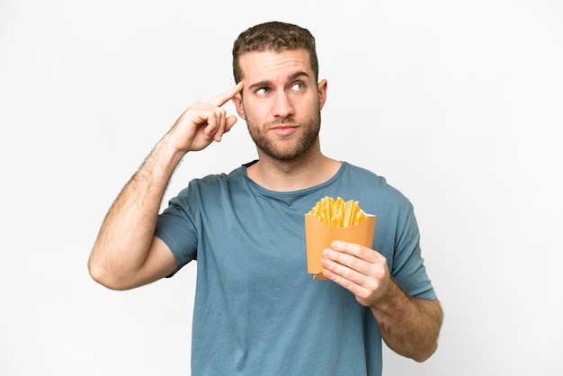 Giovane uomo biondo bello che tiene patatine fritte su sfondo bianco isolato avendo dubbi e pensando