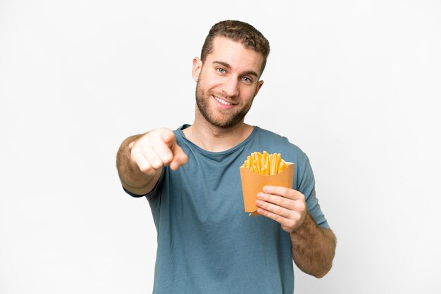 Giovane uomo biondo bello che tiene patatine fritte su sfondo bianco isolato che punta davanti con espressione felice