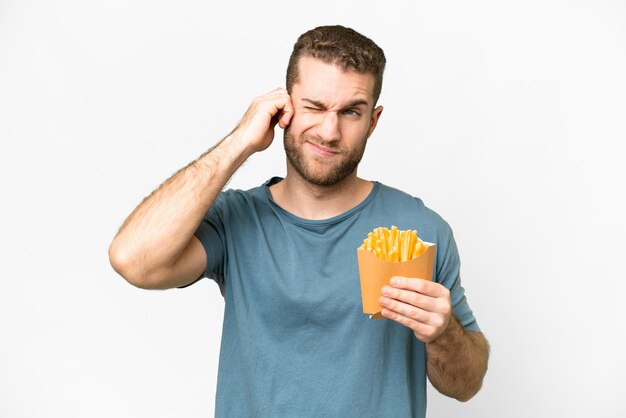 Giovane uomo biondo bello che tiene patatine fritte su sfondo bianco isolato frustrato e che copre le orecchie