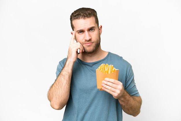 Giovane uomo biondo bello che tiene patatine fritte su sfondo bianco isolato pensando un'idea