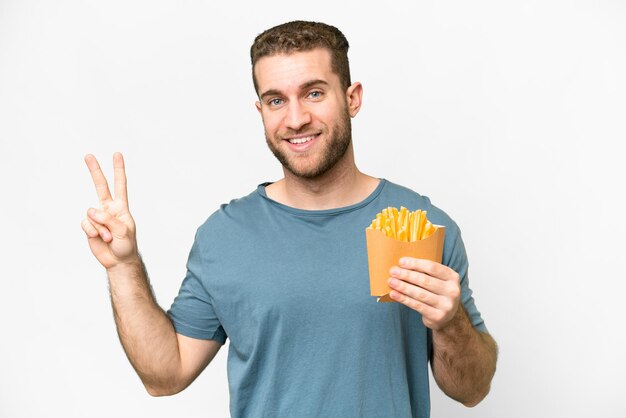 Giovane uomo biondo bello che tiene patatine fritte su sfondo bianco isolato sorridente e mostrando segno di vittoria