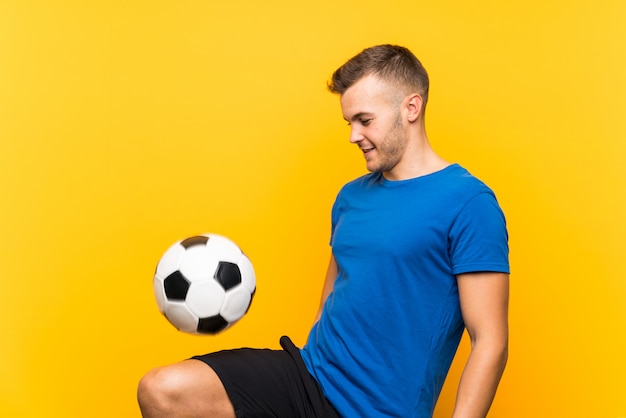 Giovane uomo biondo bello che tiene un pallone da calcio sopra fondo giallo isolato