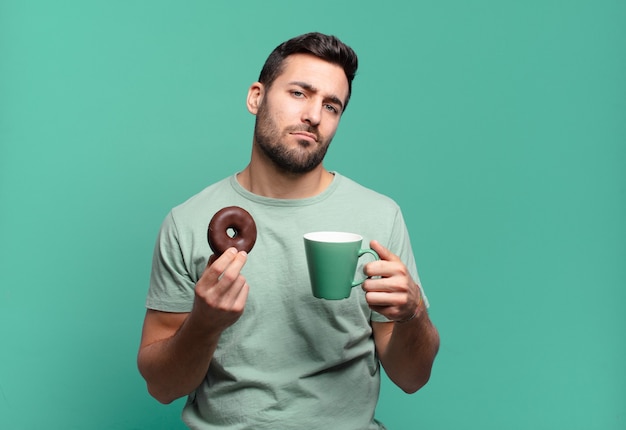 Giovane uomo biondo bello con una ciambella al cioccolato e una tazza di caffè. concetto di colazione