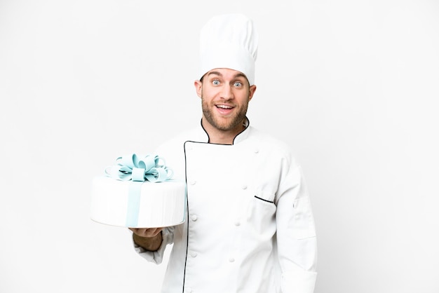 Giovane uomo biondo bello con una grande torta su sfondo bianco isolato con espressione facciale sorpresa e scioccata