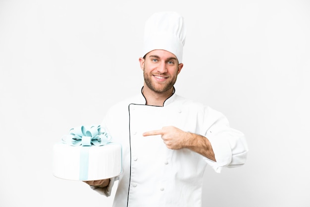 Giovane uomo biondo bello con una grande torta su sfondo bianco isolato e indicandolo